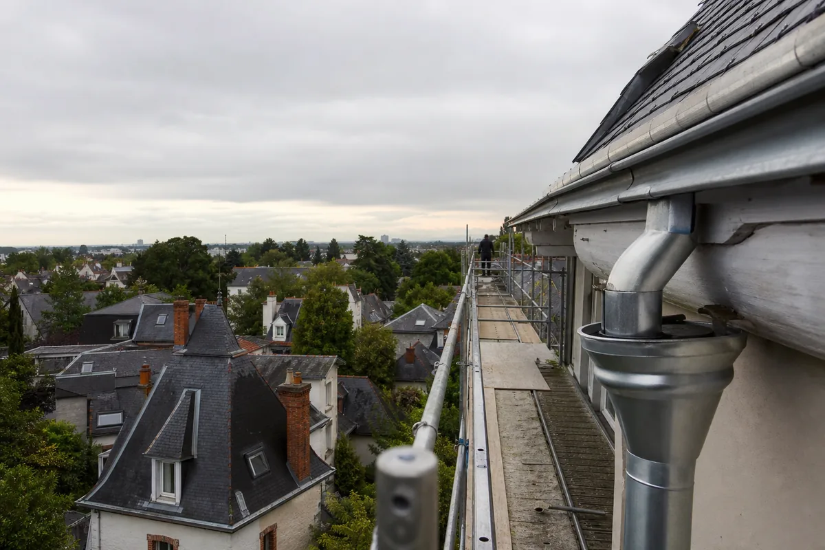 Toits d’ardoise nantais vus depuis un échafaudage, gouttière en zinc au premier plan sous un ciel gris perle